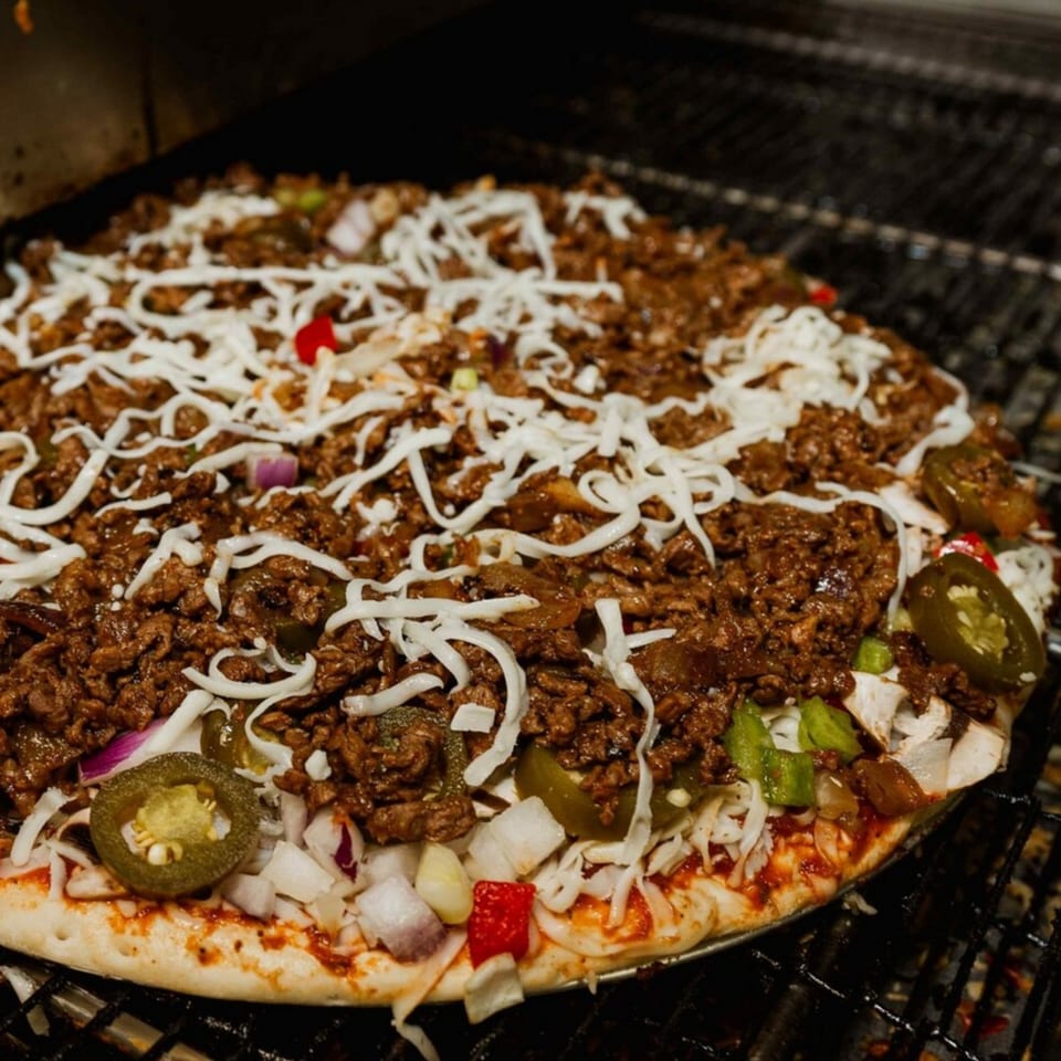 Uncooked pizza with ground meat, shredded cheese, jalapeños, and onions on a grill rack.